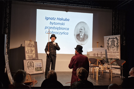 Na scenie stoi mężczyzna ubrany w strój stylizowany na dawny galowy mundur, z cylindrem na głowie i długim kijem w dłoni. Mówi do publiczności, trzymając mikrofon. Za nim wyświetlony jest slajd z napisem „Ignatz Hakuba, bytomski przedsiębiorca i dobroczyńca” oraz archiwalnym portretem mężczyzny. Po obu stronach sceny ustawiono plansze i reprodukcje starych fotografii oraz ilustracji związanych z postacią przedstawianą w prezentacji. Przed sceną siedzi publiczność, a jedna z osób w bordowym stroju i cylindrze stoi bliżej prowadzącego, tyłem do aparatu. Całość odbywa się w przyciemnionej Galerii Fermata Godula.