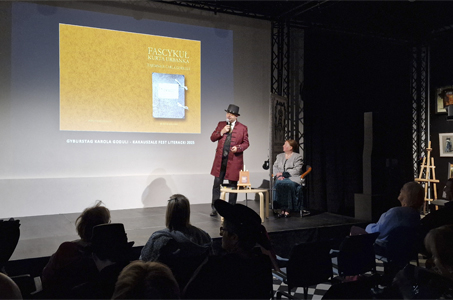 Na scenie stoi mężczyzna w bordowym fraku i cylindrze, przemawia do mikrofonu, wskazując na ekran z prezentacją zatytułowaną „Fascykuł Kurta Urbanka – Tajemnice Carla Goduli”. Obok, na krześle, siedzi kobieta w eleganckim stroju, patrząc w stronę prelegenta. Przed nią na małym stoliku leży książka. 