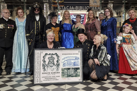 Grupowe zdjęcie osób w strojach historycznych i stylizowanych, ustawionych w holu biblioteki. W pierwszym rzędzie trzy osoby klęczą przy dużym plakacie przedstawiającym Ignatza Hakubę oraz dawne ilustracje związane z jego działalnością. Za nimi stoi kilkanaście osób w eleganckich sukniach, mundurach oraz kostiumach nawiązujących do epoki. Po prawej stronie stoi uśmiechnięta dziewczynka w tradycyjnym stroju ludowym, oparta o kobietę w kwiecistej spódnicy. W tle widać szkło, światła i szyld „STACJA BIBLIOTEKA”.