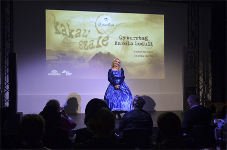 Na zdjęciu widać kobietę w długiej, niebieskiej sukni stojącą na scenie przed publicznością. Za nią znajduje się duży ekran z projekcją zawierającą tytuł wydarzenia „Gyburstag Karola Goduli” oraz informację o występie muzycznym Sylwii Szydy. Widownia siedzi w półmroku, a scena jest oświetlona jasnym światłem.