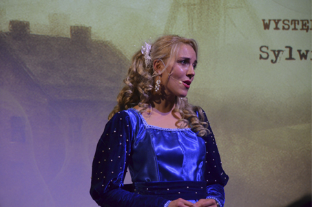 Zbliżenie na kobietę, która stoi na scenie. Ma długie, kręcone blond włosy i mikrofon nagłowny. Jest ubrana w elegancką, intensywnie niebieską suknię z błyszczącej tkaniny, zdobioną drobnymi perełkami. W tle widać rozmytą projekcję z fragmentem tekstu z wydarzenia. Kobieta wygląda, jakby śpiewała.