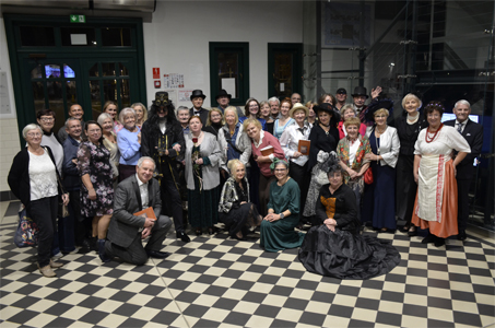 Duża grupa osób pozuje wspólnie do zdjęcia w jasnej, przestronnej hali z przeszklonymi drzwiami i oknami w tle. Część osób ubrana jest w stroje stylizowane na dawne epoki – między innymi suknie, kapelusze, cylindry, stroje steampunkowe i kostiumy historyczne. Inni uczestnicy pozują w codziennych ubraniach. Wszyscy stoją lub klęczą na czarno-białe podłodze na wzór szachownicy. Grupa uśmiecha się i patrzy w stronę aparatu.