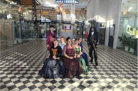 Grupa siedmiu osób w strojach stylizowanych na XIX-wieczne, pozuje na holu biblioteki. Pięć kobiet w długich, eleganckich sukniach w odcieniach granatu, zieleni, czerwieni i czerni siedzi na ławce na środku pomieszczenia. Po lewej stoi mężczyzna w bordowym fraku i cylindrze, trzymający laskę; po prawej mężczyzna w czarnym kostiumie steampunkowym z długimi włosami i ozdobnym kapeluszem. W tle widać regały z książkami oraz przeszklone pomieszczenia biblioteki.