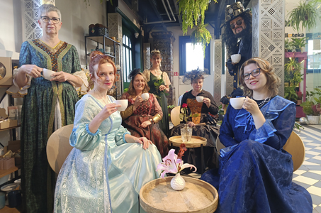 Grupa siedmiu osób w eleganckich historycznych strojach pozuje we wnętrzu pełnym roślin i dekoracji. Sześć osób — pięć kobiet i jeden mężczyzna w stroju steampunkowym — trzyma filiżanki z herbatą lub kawą, siedząc przy okrągłych stolikach lub stojąc za nimi. Kobiety mają długie suknie w kolorach zieleni, błękitu, granatu, brązu i czerni, a jedna z nich nosi ozdobny kapelusz. Mężczyzna w cylindrze z okularami i ozdobami steampunkowymi pochyla się zza dekoracyjnej ścianki, również trzymając filiżankę. W tle widać wiszące paprocie, regały oraz elementy wystroju w stylu industrialno-artystycznym.